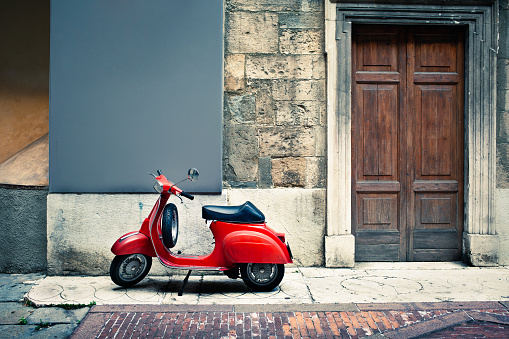 A vintage red Italian scooter stands on a sidewalk beside an old-fashioned wooden door.  The wall and sidewalk are clearly old and are patched with various materials including stone and brick.  The scooter is clean and shiny looking, with a glossy black seat.
