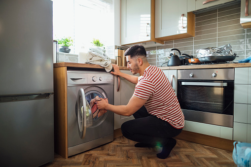 Man crouching down to put washing into his washing machine in his kitchen.  He is in the Northeast of England.
