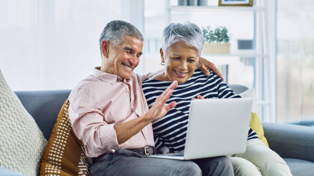 It's like their loved ones are in their living room Shot of a senior couple waving while using a laptop together on the sofa at home free video chat with women stock pictures, royalty-free photos & images