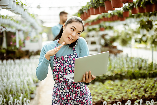 Happy Asian businesswoman talking on a cellphone while standing in her greenhouse. Joyful female florist calling on smartphone at work. Own business concept