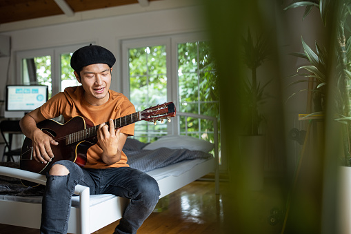 Japanese man sitting on bed and enjoying while playing acoustic guitar in bedroom at home
