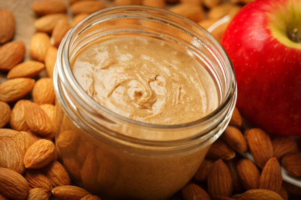 Jar Of Almond Butter Surronded By Almonds And An Apple On open jar of almond butter is surrounded by a bed of almonds and a red apple. almond butter stock pictures, royalty-free photos & images