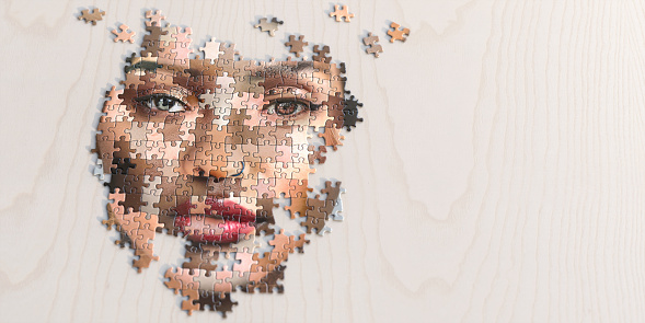 A close up image of a partly assembled jigsaw puzzle of a generic female face made of many different ethnicities and skin colours. The puzzle is on a light plain wooden surface, and shot with shallow depth of field with focus on the eyes. With copy space.