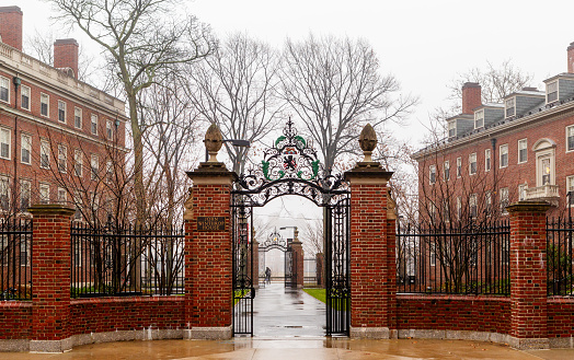 Cambridge, Massachusetts, USA - December 11, 2021: The John Winthrop House and gate in Harvard Yard on a rainy day. Winthrop is one of twelve undergraduate residential Houses at Harvard University. It is home to approximately 400 upperclass undergraduates. Winthrop house consists of two buildings, Standish Hall and Gore Hall built in 1912. In 1931 they were joined as John Winthrop House. The House crest atop the gate shows a lion on a shield with chevrons in the background.