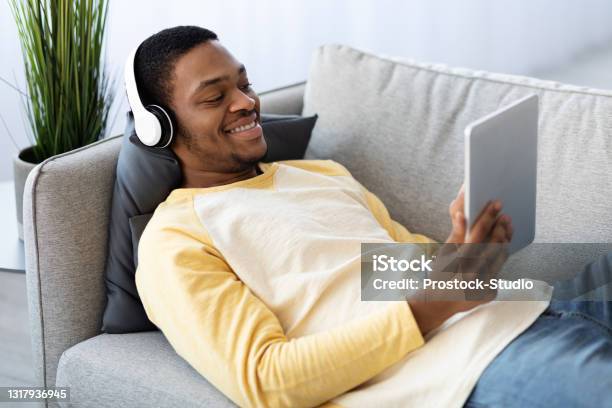 Joyful black guy laying on couch, watching movies on pad
