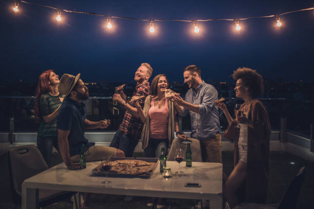 Joyful friends having a dinner party during the night on a terrace. Large group of happy friends having fun while eating pizza and drinking alcohol during night party on a penthouse terrace. roof garden stock pictures, royalty-free photos & images