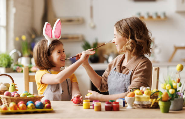 Joyful happy family mom and daughter having fun and fooling around while decorating Easter eggs Funny photo of joyful happy family mom and little daughter having fun and fooling around while decorating Easter eggs together for spring holidays, sitting at table in cozy kitchen. easter sunday stock pictures, royalty-free photos & images