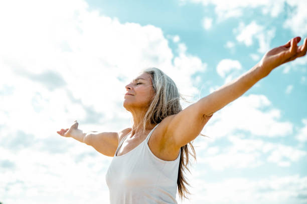 Joyful senior woman enjoying freedom standing with open arms and a happy smile looking up towards the sky - People and happiness concept stock photo