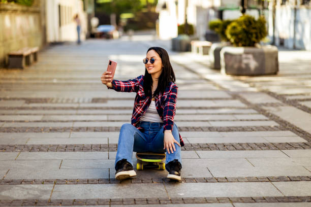 vrolijke skater gekleed in modieuze streetwear klikken inhoud selfie tijdens vrije tijd in de stad, oprecht hipster meisje zittend aan longboard en het creëren van invloed vlog voor het delen met sociale netwerken - streetwear stockfoto's en -beelden