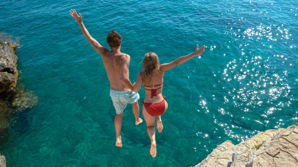 Joyful tourist couple decides to jump off a rocky cliff and dive into sea. stock photo
