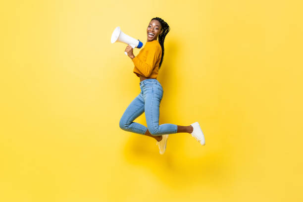 Joyful young African American woman holding megaphone jumping and making announcement in isolated studio yellow background stock photo