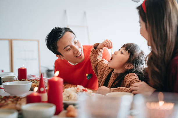 Joyful young Asian family spending time celebrates Christmas and enjoying Christmas party together at home while little girl dangles spaghetti into her mouth Joyful young Asian family spending time celebrates Christmas and enjoying Christmas party together at home while little girl dangles spaghetti into her mouth asian family eating together stock pictures, royalty-free photos & images