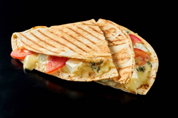 Juicy Mexican quesadilla with mozzarella, camembert, blue cheese and tomatoes. Isolated on black background. Street food. Close up, selective focus stock photo