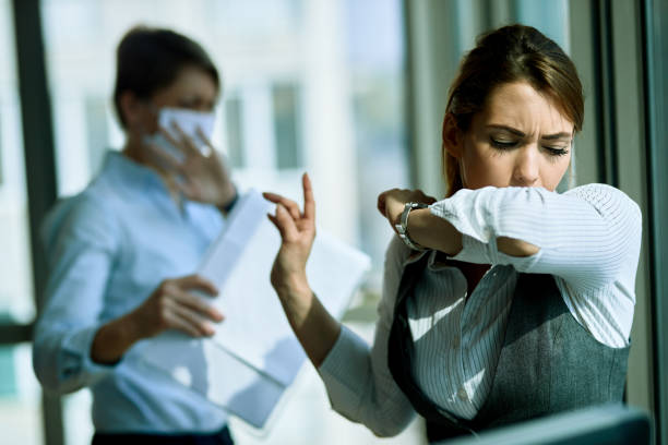 Just a second please, this cough is catching me! Young businesswoman coughing into elbow in the office. Her coworker is in the background. sneezing stock pictures, royalty-free photos & images