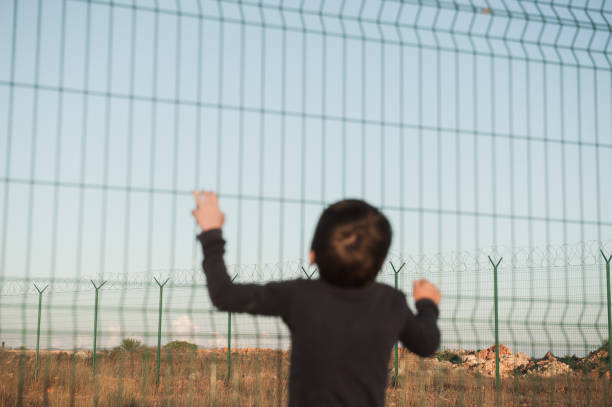 juvenile delinquent little refugee ilegal immigrant kid in displaced persons camp with high fence with barbed razor wire with hope of freedom in Mexico USA Europe Turkey Syria juvenile delinquent little refugee kid in displaced persons camp with high fence with barbed razor wire with hope of freedom geographical border stock pictures, royalty-free photos & images