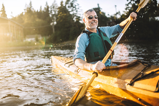A mature adult man glides through the cool waters of Gig Harbor, Washington, a small bay in the Puget sound, paddling in a classic wood kayak.  The evening sun shines from behind, a smile on the mans face as he takes in the beauty of the great outdoors.  Horizontal image with copy space.