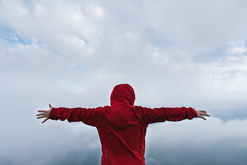Back view of a kid with extended arms in front of a dramatic sky and wearing a colorful red raincoat. Feeling free and flying sensation concept.