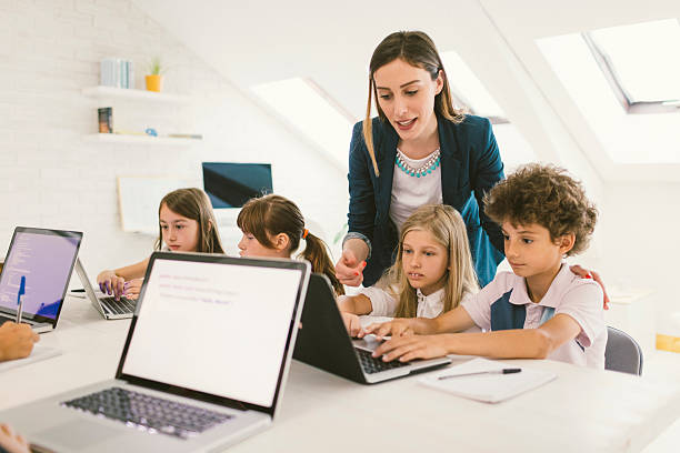 Kids Coding In School Group of children at coding class. They are sitting by the desk and using laptops. Their female teacher standing beside them and helping with their assignment. programming languages to teach kids stock pictures, royalty-free photos & images