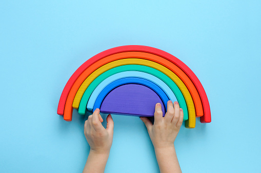 Kids hands playing with wooden toy rainbow on blue background. Top view, flat lay.