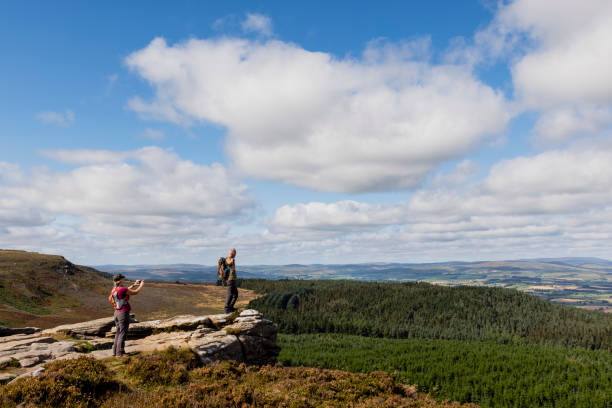 King of the Mountain A woman and her brother on a hike in Rothbury, Northumberland. They are taking a break from walking and the woman is taking a photo with her mobile phone of the man, who is standing on the edge of a cliff with his arms out. rothbury northumberland stock pictures, royalty-free photos & images