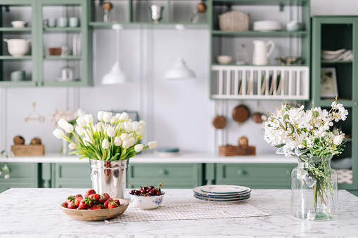 Kitchen island with marble surface, small bowl of cherries with bigger wooden bowl filled with strawberry, flowers in bucket and glass jar, green vintage cupboards full of different devices