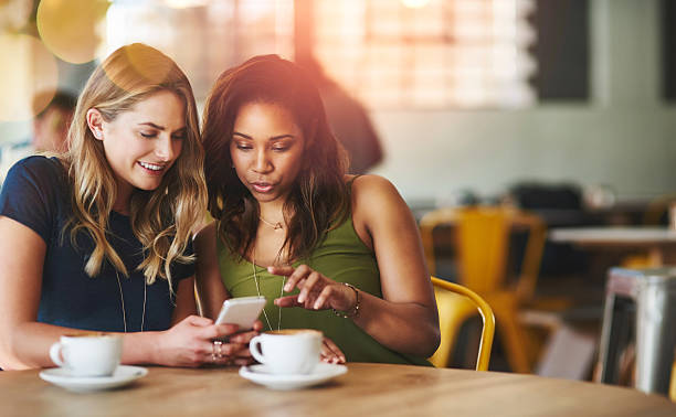 I know exactly what you can tell him! Cropped shot of girlfriends sharing something on a cellphone while sitting in a cafe female friendship photos stock pictures, royalty-free photos & images