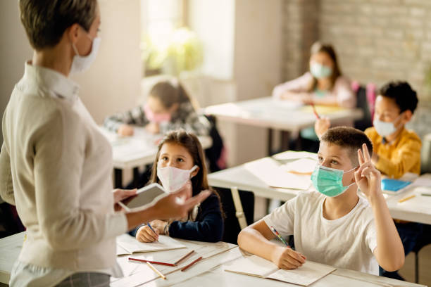 I know the answer! Schoolboy with face mask raising hand to answer the question during a class in the classroom. elementary school building stock pictures, royalty-free photos & images