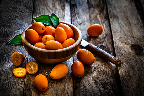 Kumquats shot on rustic wooden table Close up view of fresh ripe organic kumquats shot on rustic wooden table. The kumquats are in a wooden bowl and some are scattered on the table around the bowl. A single kumquat is sliced and a kitchen knife complete the composition. Predominant colors are orange and brown. High resolution 42Mp studio digital capture taken with Sony A7rii and Sony FE 90mm f2.8 macro G OSS lens kumquat stock pictures, royalty-free photos & images