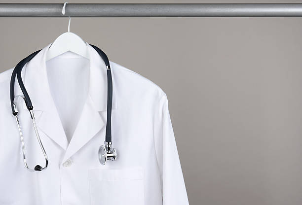 Lab Coat with Stethoscope on Hanger A doctor's lab coat and stethoscope on hanger against a gray background. Closeup on a white hanger with a gray background in horizontal format with copy space. lab coat stock pictures, royalty-free photos & images