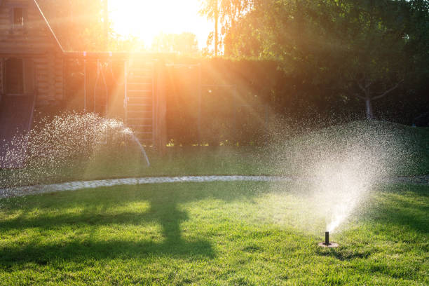 landschap automatische tuin watersysteem met verschillende sprinklers geïnstalleerd onder grasmat. landschapsontwerp met gazonheuvels en fruittuin die met slimme autonome sproeiers bij zonsondergangtijd wordt geïrrigeerd - irrigatiesysteem stockfoto's en -beelden
