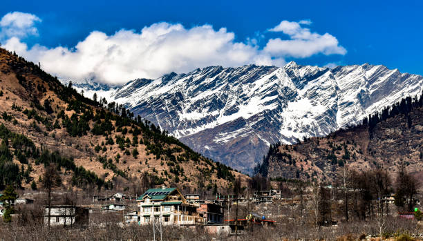 Landscape of snow covered mountains of Himalayas in Background, and in foreground, mountains with deciduous forests with beautiful Hamlet or Village in valley on bright sunny day with cloudy blue sky. Beautiful Snow covered mountains of Great Indian Himalayan Ranges. The mountain in the background is covered in Ice for being situated at high Altitude. Human Settlement could be seen in the foreground. Clear blue sky and a bright white cloud is adding beauty to the photo. shimla stock pictures, royalty-free photos & images