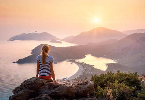 Young woman sitting on the top of rock and looking at the seashore and mountains at colorful sunset in summer. Landscape with girl, sea, mountain ridges and orange sky with sun. Oludeniz, Turkey.