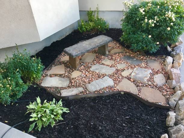 Landscaped Flowerbed with Bushes A newly landscaped flowerbed with flat rocks, orange pebbles, a rose bush, Snapdragon flowers, dark mulch, and a stone bench. mulch stock pictures, royalty-free photos & images