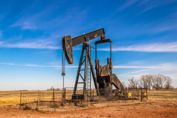 Large old rusty oil well pump jack surrounded by cattle panel fence out in field with very blue sky in winter. stock photo