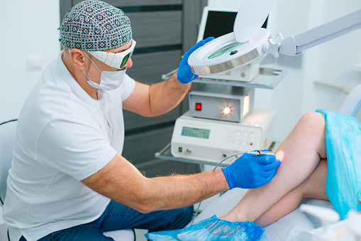 Concentrated dermatologist using professional equipment and a magnifying glass while treating the veins on a leg of his patient