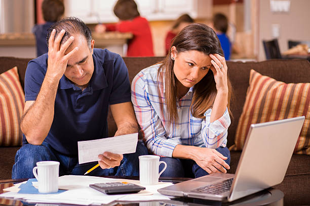 Latin descent couple paying monthly bills at home.  Frustration. Latin descent man and woman struggle to pay their monthly bills.  They are calculating expenses versus budget income and are upset.  Many invoices, laptop on living room coffee table.  Children in kitchen background.  Mid-adult age couple.  Frustration among middle-class people.   Great imagery for election season:  home finances, recession, past due bills, mortgage, debt, stress, worry, taxes. foreclosure stock pictures, royalty-free photos & images