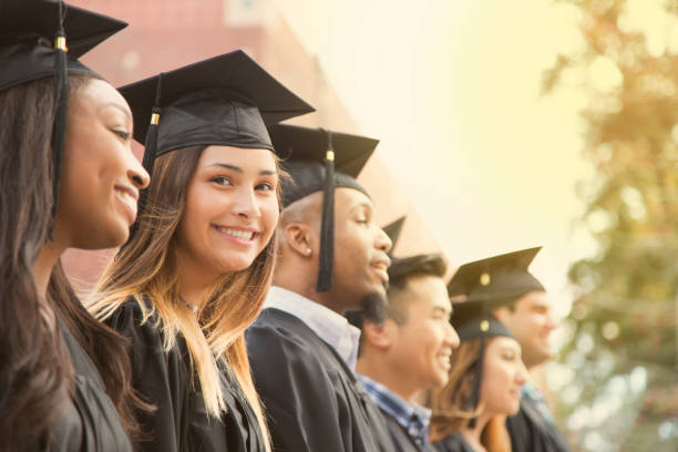 Latin descent female college student graduation on campus. Latin descent girl waits in line during college graduation ceremony. She looks at camera with a big smile as she wears a black cap and gown. universities in usa stock pictures, royalty-free photos & images