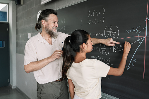 Latin Female Teenager Student doing Maths Geometry Exercises on Blackboard with help of friendly professor in High School