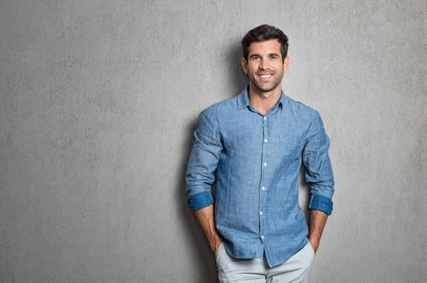 Latin man standing Portrait of a handsome young man smiling against grey background with copy space. Smiling latin guy with hands in pocket in blue shirt standing and leaning on wall. Successful hispanic man looking at camera. handsome people stock pictures, royalty-free photos & images