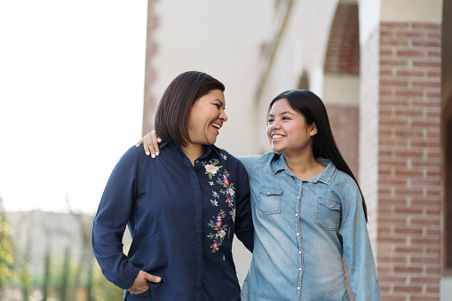 A latin mother and her teenage daughter standing together, embracing and smiling.