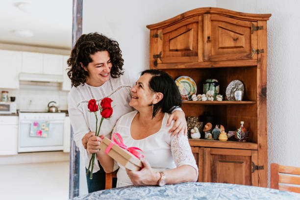 latin young woman and her mother middle age with flowers and gift box at home celebrating Happy mother's day in Mexico city stock photo