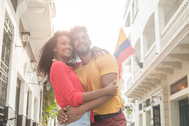 Latino couple hugging in downtown Cartagena Latin couple made up of black curly-haired woman in brown skin, red blouse and jean short that is hugging her boyfriend Latin man with curly hair and yellow sweater who is also hugging her while they know the historic center of the city of Cartagena where We observe old house with flags of Colombia colombian ethnicity stock pictures, royalty-free photos & images