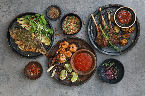 Brazilian coconut shrimp kebabs. Barbecued sea bream with grilled vegetables. Brazilian grilled lamb chops. Flat lay top-down composition on concrete background.