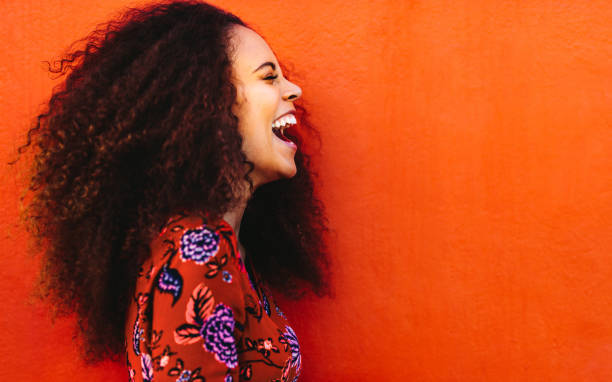Laughing african young woman with curly hair Side view of cheerful woman with curly hair standing against orange background. Close up of laughing african young woman. laughing stock pictures, royalty-free photos & images