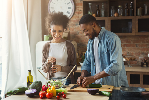 Laughing black couple preparing healthy salad together in loft kitchen. Young family cooking dinner