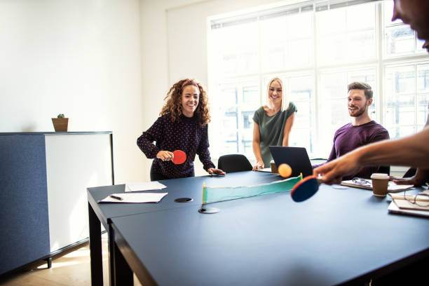 Laughing colleagues playing table tennis together on a boardroom table Laughing group of young designers playing table tennis on a boardroom table during a break from an office meeting table tennis stock pictures, royalty-free photos & images
