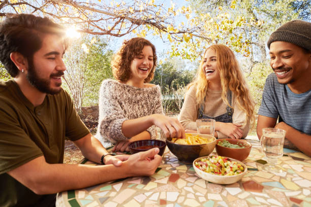 Laughing friends eating nachos togegher on an outdoor patio Group of diverse young friends laughing and eating snacks outdoors at a table on a farm dipping sauce stock pictures, royalty-free photos & images