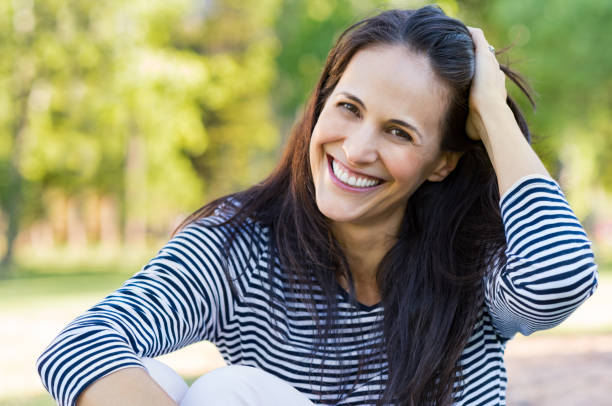 Laughing mid woman at park Happy latin woman having fun at park while touching hair. Hispanic woman enjoying holiday while sitting on grass in park. Portrait of smiling mature woman relaxing and looking at camera. mid adult women stock pictures, royalty-free photos & images