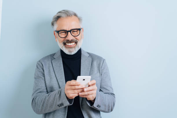 Laughing middle-aged man with smartphone Bearded middle-aged man with grey hair, holding smartphone, looking at camera and laughing. Half-length front portrait against blue wall background with copy space 50 54 years stock pictures, royalty-free photos & images
