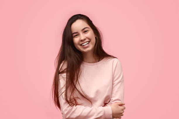 Laughing teenager with folded arms Happy teen girl with long hair laughing and embracing torso while standing against pink background adolescence stock pictures, royalty-free photos & images